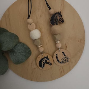 May include: Two wooden horse-themed necklaces with black cords. One necklace features a horse head charm, a white pom pom, and a horseshoe charm with flowers. The other necklace has a horse head charm, a heart charm, and a horse head charm.