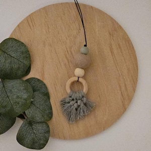 Peut inclure: Un collier en macramé gris et beige avec des perles en bois et un anneau en bois. Le collier est suspendu à une surface en bois.