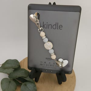 May include: A grey Kindle e-reader with a beaded charm. The charm has a silver clasp, heart-shaped details, and a string of light-coloured beads. The word "kindle" is printed on the device.
