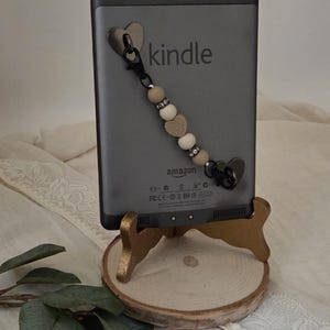 May include: A grey Kindle e-reader with a beaded charm featuring heart-shaped and round beads. The Kindle is displayed on a small, gold-painted wooden stand with a natural wood slice base. The word "kindle" is printed on the device.