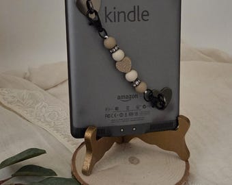 May include: A gray Kindle e-reader with a beaded charm featuring heart-shaped and round beads. The Kindle is displayed on a small, gold-painted wooden stand with a natural wood slice base. The word "kindle" is printed on the device.