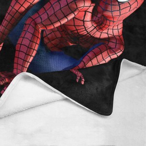 Op de afbeelding: Een zwart en rood fleece deken met een rood en blauw Spiderman ontwerp. Het deken is dubbelgevouwen, waardoor de witte fleece voering zichtbaar is.