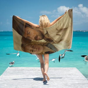 May include: A beach towel with a photo of a man and woman on it. The towel is brown and beige with a faded look. The man and woman are wearing casual clothing. The towel is being held by a person on a beach.