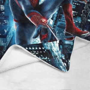 Pu&ograve; includere: Una coperta in pile bianca con un'immagine blu e rossa di Spiderman che si dondola su uno skyline urbano.