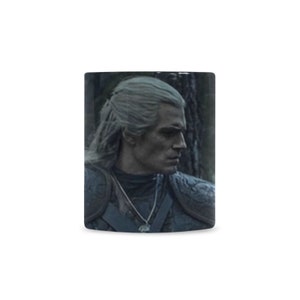 Op de afbeelding: Een zwart-witte mok met een foto van een man met wit haar en een baard. Hij draagt ​​een donker leren jack en een ketting. De achtergrond is een donker bos.