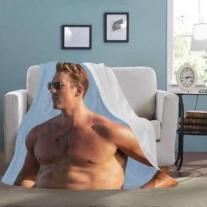 May include: A blue and white throw blanket featuring a photograph of a shirtless man wearing sunglasses on a beach.