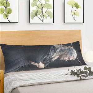 May include: A body pillow featuring a black and white image of a man with long white hair. The man is wearing a black shirt and looking down. The pillow is on a white bed with a wooden headboard.