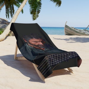 May include: A black beach towel with a graphic of a man's face and a guitar. The towel is draped over a wooden beach chair.