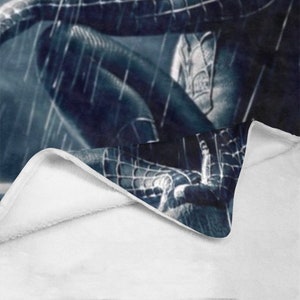 May include: A black and blue fleece blanket with a superhero character design. The blanket is folded in half, showing the white fleece lining.