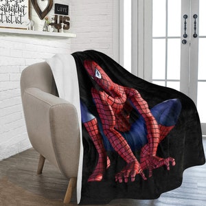 Op de afbeelding: Een zwarte en rode fleece deken met een afbeelding van Spiderman in een hurkende houding. De deken is over een stoel gelegd.