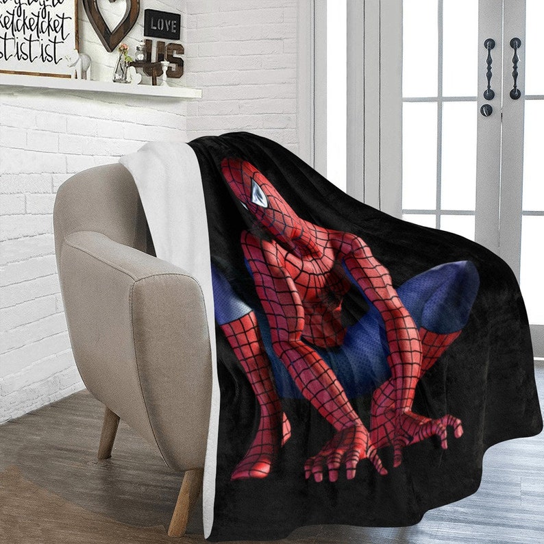Op de afbeelding: Een zwarte en rode fleece deken met een afbeelding van Spiderman in een hurkende houding. De deken is over een stoel gelegd.