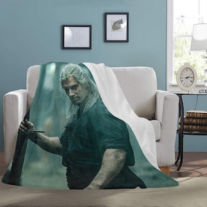 May include: A white throw blanket featuring a printed image of a man with long white hair holding a sword. The man is wearing a dark green shirt and black trousers. The background is a blurred green and blue.
