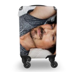 May include: Rolling suitcase with a photographic print of a man's face. The man has dark hair, a mustache, and a beard. He is wearing a grey shirt and is lying down. The suitcase has black wheels and a black handle.