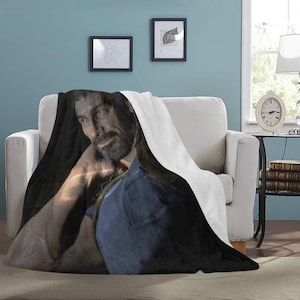 May include: A black and white throw blanket with a man's face printed on it. The man has a beard and is wearing a blue shirt. The blanket is folded and sitting on a white couch.