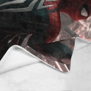 May include: A white fleece blanket with a red and black superhero design printed on one side. The design features a superhero in a red and black suit with a spider web pattern.
