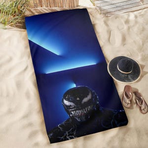 May include: A blue beach towel featuring a graphic of the Venom character. The towel is on a sandy surface, with a black and white sun hat and a pair of brown flip-flops.