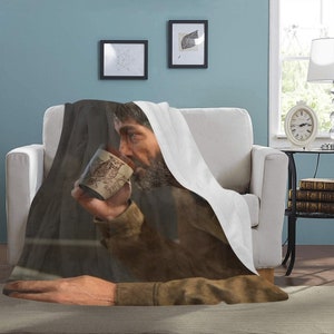 May include: A soft, white throw blanket with a brown and black image of a man drinking from a mug. The mug has a brown owl design. The man is wearing a brown jacket and has a beard.