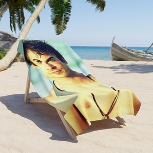 May include: A beach towel with a photo of a man with blue eyes and a necklace. The towel is folded and placed on a wooden beach chair. The chair is on a sandy beach with palm trees in the background.