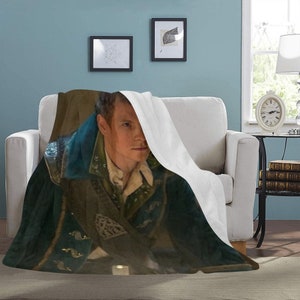 Puede incluir: Un hombre con una chaqueta azul y verde, una camisa blanca y un cuello blanco está parcialmente oculto bajo una manta blanca. El hombre mira a la cámara con una expresión seria. La manta está en un sofá con una pared azul claro en el fondo.