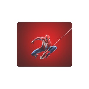 以下が含まれることがあります： 赤い背景に、スパイダーマンがダイナミックなポーズをとっている長方形のマウスパッド。スーパーヒーローは、胸に白いクモのエンブレムが付いた、彼のクラシックな赤と青のスーツで描かれています。