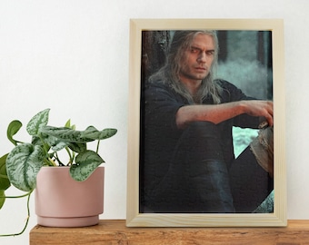 Op de afbeelding: Een ingelijste puzzel met een man met lang, grijs haar die in een bosrijke omgeving zit. De man draagt een zwart shirt en broek. De puzzel is gemaakt van hout en heeft een natuurlijke afwerking.