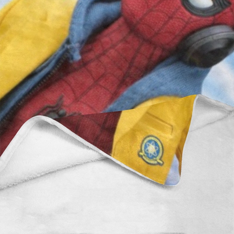 May include: A white fleece blanket with a red and blue superhero character printed on it. The character is wearing a yellow jacket with a blue and white logo on the front. The blanket is folded in half and the character is visible on the top half of the blanket.