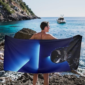 May include: A dark blue beach towel with a graphic design of a black creature. The towel is held up by a person on a rocky beach with the ocean and a boat in the background.