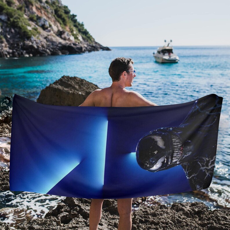 May include: A dark blue beach towel with a graphic design of a black creature. The towel is held up by a person on a rocky beach with the ocean and a boat in the background.