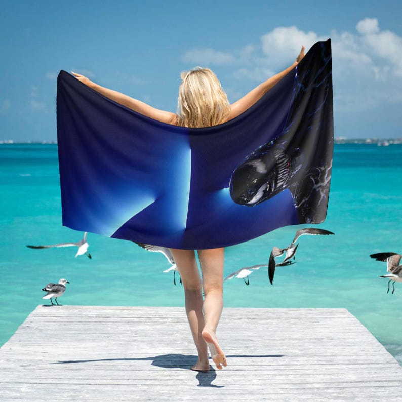 May include: A dark blue beach towel with a whale design. The towel is held up by a person on a wooden dock, with a turquoise ocean and seagulls in the background. The whale design is black and white.