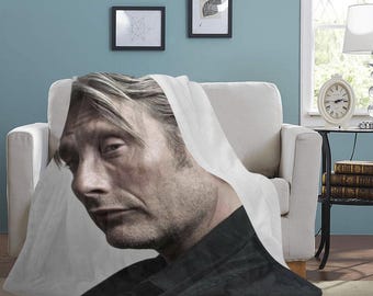 May include: A white throw blanket with a man's face printed on it. The man has short, light brown hair and is wearing a black shirt. The blanket is folded over a white couch.