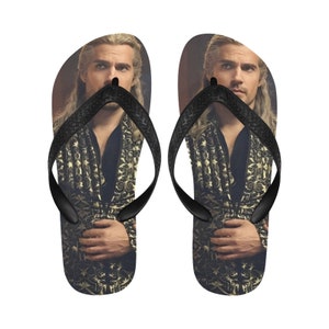 May include: A pair of black flip-flops with a photo of a man with long blonde hair wearing a gold and black patterned jacket.