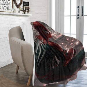 May include: A red and black fleece throw blanket with a graphic of Spider-Man in a city setting. The blanket is draped over a light brown armchair.