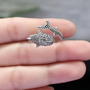 K&ouml;nnte beinhalten: Ein silberner Ring in Form eines Walhais. Der Ring hat ein detailliertes Design mit kleinen, dunklen Akzenten. Der Schwanz des Hais umschlingt den Finger und schafft ein einzigartiges und auff&auml;lliges Schmuckst&uuml;ck.