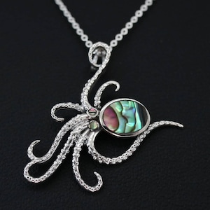 Op de afbeelding: Een zilveren octopus hanger ketting met een ovaal abalone schelp centrum. De hanger heeft gedetailleerde tentakels en een delicate ketting. De abalone schelp toont iriserende kleuren van roze, groen en blauw. De ketting wordt gepresenteerd tegen een zwarte achtergrond.