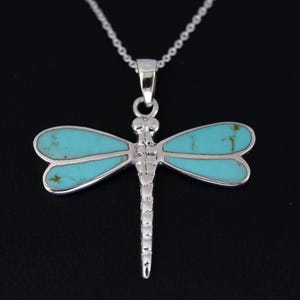May include: A silver dragonfly pendant necklace with turquoise-colored wings. The dragonfly has a detailed body and is suspended from a delicate chain. The necklace is set against a black background.