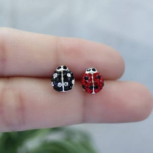 May include: A pair of ladybug stud earrings. One earring is black with clear crystal accents, the other is red with black accents. The earrings are small and detailed, with a silver-colored metal base.