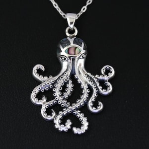 Op de afbeelding: Een zilveren ketting met een octopus hanger. De octopus heeft gedetailleerde tentakels en een kop ingelegd met iriserende abalone schelp. De ketting is ook zilver. De hanger wordt gepresenteerd tegen een zwarte achtergrond.