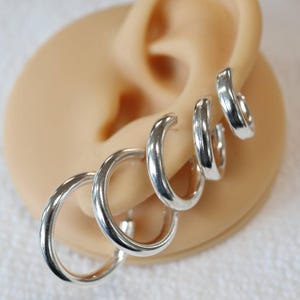 May include: Four silver hoop earrings of different sizes, stacked on a model ear.