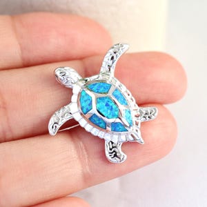 May include: A silver-colored turtle brooch with a bright blue center. The brooch is held in a hand, with a small plant in the background. The turtle design has intricate details.