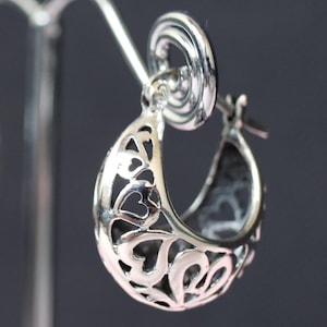 May include: A pair of silver hoop earrings with a delicate, intricate design featuring cut-out hearts. The earrings are displayed on a silver stand.