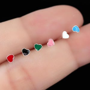 Könnte beinhalten: Eine Sammlung von sieben kleinen herzförmigen Ohrsteckern in verschiedenen Farben, darunter Rot, Schwarz, Grün, Rosa, Weiß und Blau. Die Ohrringe werden auf einem Finger vor dunklem Hintergrund präsentiert. Diese Ohrringe sind ein süßes Accessoire.