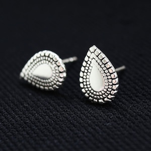 May include: A pair of silver teardrop stud earrings. Each earring has a smooth, central teardrop shape surrounded by a detailed, textured border. The earrings are shown against a black background, emphasising their metallic sheen.