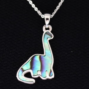 Könnte beinhalten: Eine silberne Halskette mit einem Anhänger in Dinosaurierform. Der Anhänger ist mit schillerndem Abalone-Muschel gefüllt, das Blau-, Grün- und Lilatöne aufweist. Die Halskette hat eine feine Kette und einen Karabinerverschluss.