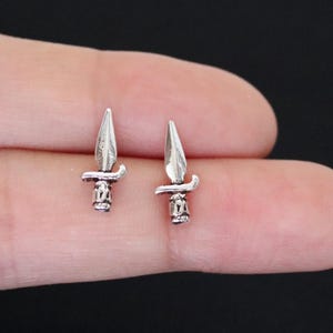 Op de afbeelding: Een paar zilveren dolk oorringen. De oorringen zijn klein en delicaat, met een scherpe punt en een gedetailleerd handvat.