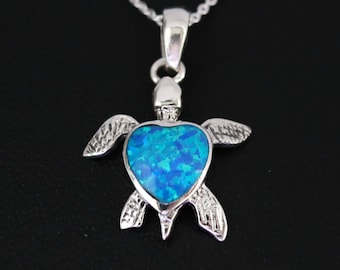 May include: A silver pendant necklace with a turtle design. The turtle's body is a heart shape filled with blue opal. The necklace has a fine chain and is set against a black background.