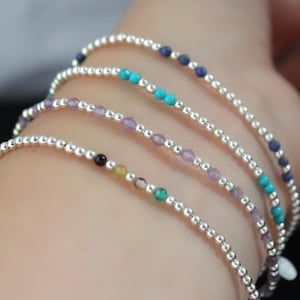 Op de afbeelding: Vier delicate armbanden met zilveren kralen en kleurrijke edelstenen. De armbanden hebben turquoise, paarse en blauwe kralen. De armbanden worden om een pols gedragen.