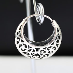 May include: A silver hoop earring with an openwork design. The earring is crescent-shaped, with intricate cut-out patterns. The metal has a polished finish, reflecting light. The earring is suspended on a silver display stand against a black and white background.