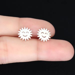 May include: A pair of small, silver-colored stud earrings. Each earring is shaped like a smiling sun with spiky rays. The earrings are held between fingers against a dark background.