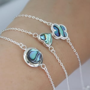 Könnte beinhalten: Drei silberne Armbänder mit Abalone-Muschel-Anhängern in runden, herzförmigen und Kleeblatt-Formen. Die irisierenden blau-grünen Muscheln sind in filigrane Silberfassungen gefasst und durch feine Kettenglieder verbunden. Die Armbänder werden an einem Handgelenk gezeigt.