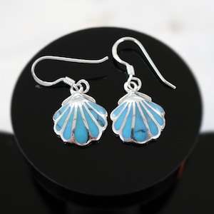 Puede incluir: Un par de pendientes de plata con forma de concha marina con incrustaciones de turquesa. Los pendientes presentan un diseño detallado de concha con una combinación de colores azul brillante y plata. Los pendientes se muestran sobre una superficie negra.